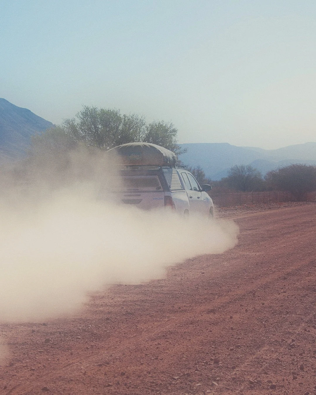 SUV mit Dachzelt fährt auf unbefestigter Straße in Namibia, aufgewirbelter Staub, bergige Landschaft.