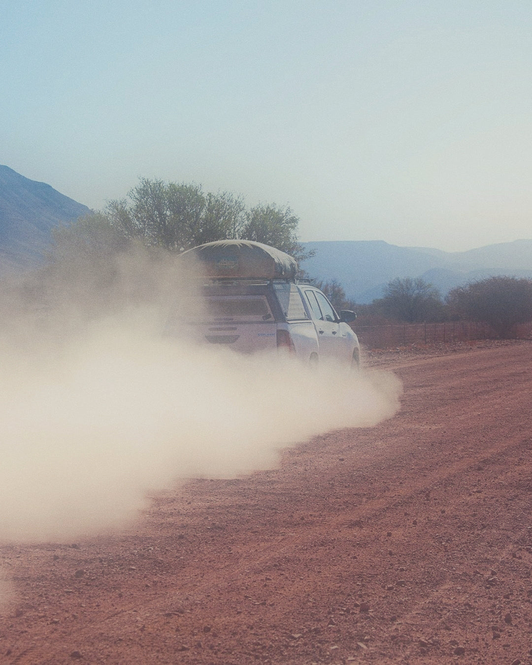 SUV con tenda sul tetto viaggia su strada sterrata in Namibia, polvere sollevata, paesaggio montuoso.